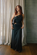 Woman in a black one-shoulder dress standing in a room with curtains and wooden floor.