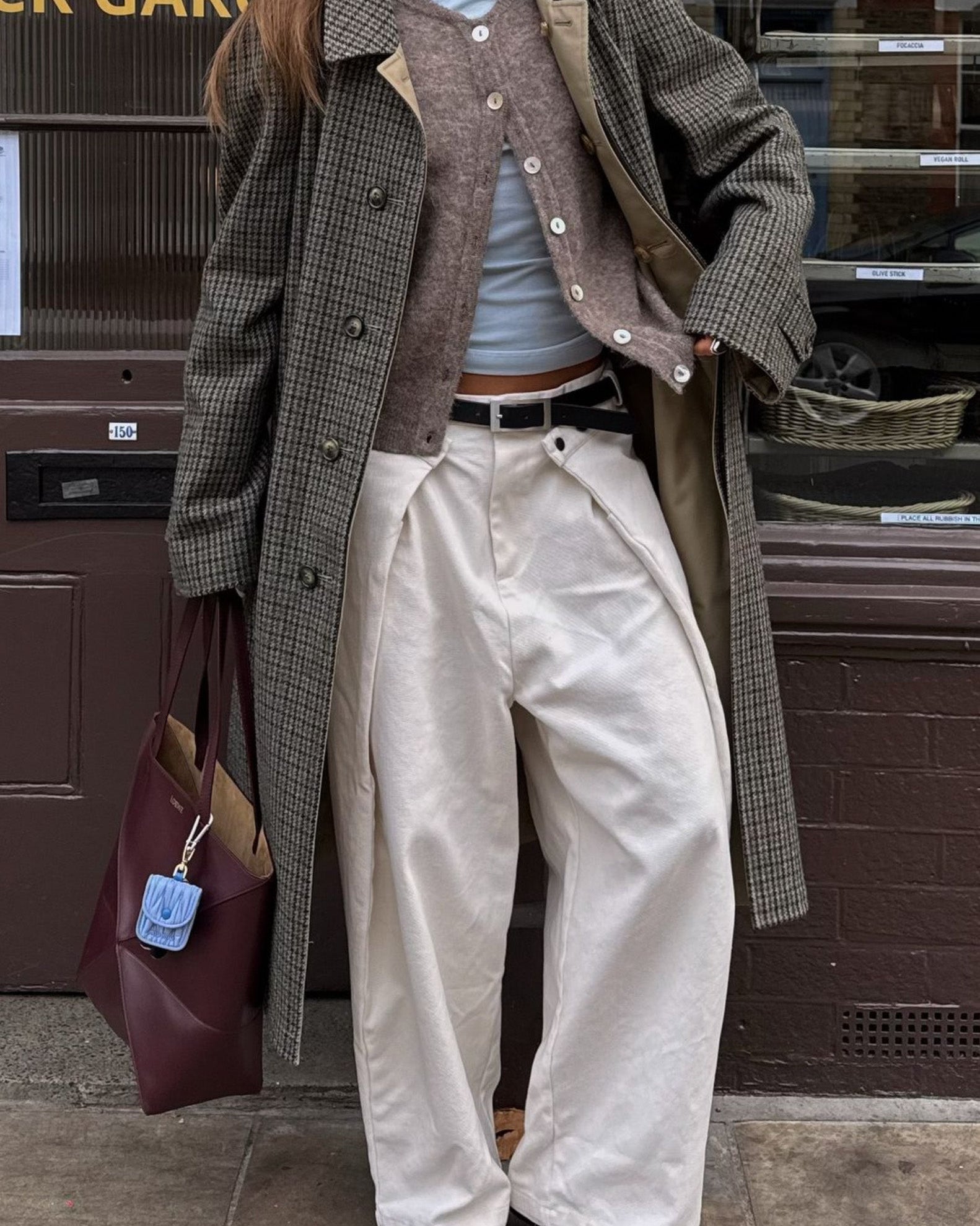 Person wearing a long coat, white pants, and holding a maroon bag with a blue tag.